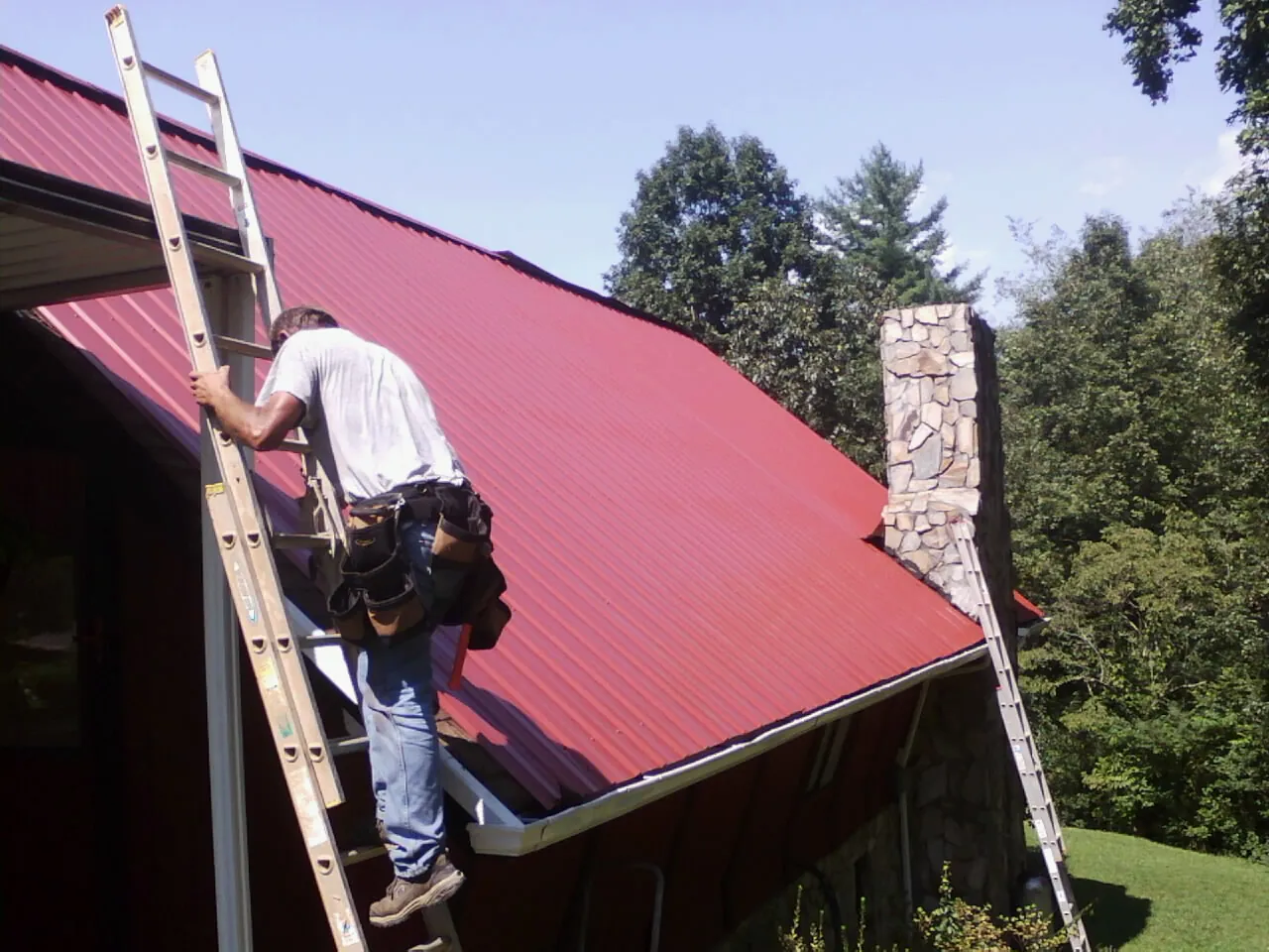 Roofer on a ladder beside a red metal roof and stone chimney — Soffit & Fascia Repair by Roofing Brothers in Brooklet